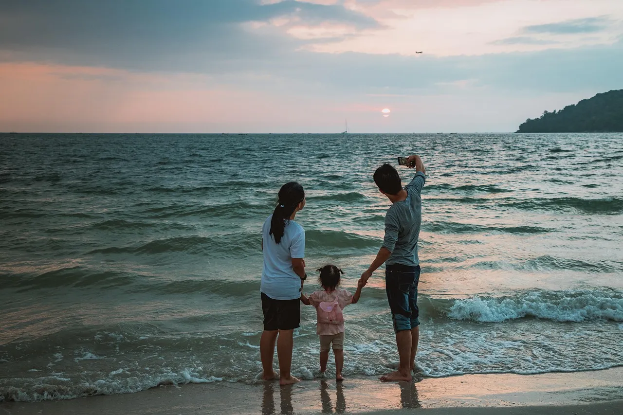 A family at the edge of the sea at sunset, the father holding up a phone to take a photo